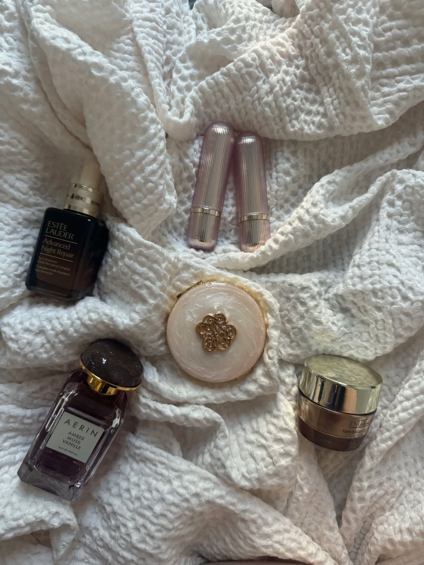 Flat lay of Estée Lauder and AERIN beauty products on white textured fabric